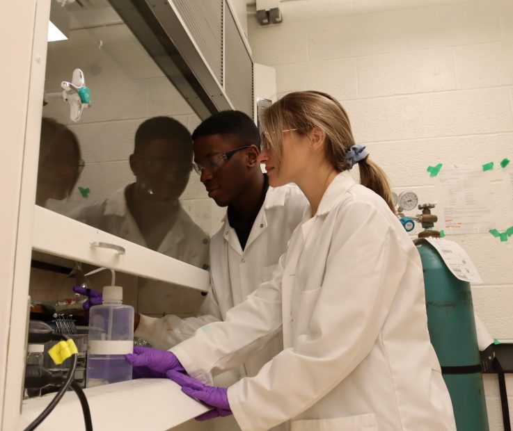 Runako Gentles and Nicolette Bugher wearing white lab coats and safety glasses measuring a solution inside a glass instrument in the Plata Lab. 